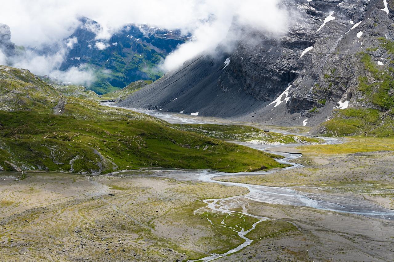 A stunning alpine landscape featuring lush green meadows, rocky terrain, and winding streams cutting through the valley. Towering grey cliffs rise in the background, partially shrouded by low-hanging clouds.