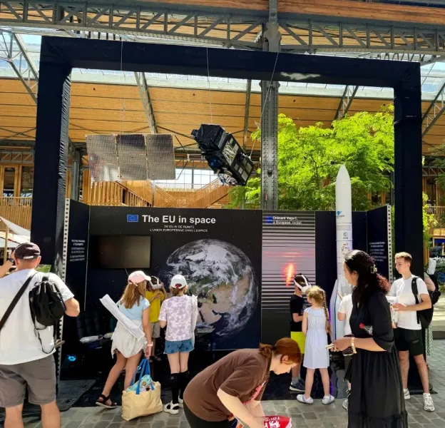 A large exhibition stand titled "The EU in space" displays a Copernicus satellite model suspended overhead, an Earth Observation image, and a Ariane 6 rocket model. Several adults and children engage with the exhibit, including one child using a virtual reality headset.