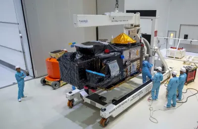 Technicians in blue coveralls and hard hats working on a black foil-wrapped satellite mounted on a transport frame in a clean room.