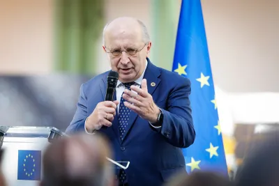 A man in a blue suit and patterned tie speaks into a handheld microphone, gesturing with his open hand, at a transparent lectern bearing the EU emblem, with a European Union flag behind him.