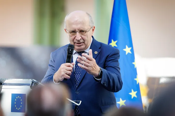 A man in a blue suit and patterned tie speaks into a handheld microphone, gesturing with his open hand, at a transparent lectern bearing the EU emblem, with a European Union flag behind him.