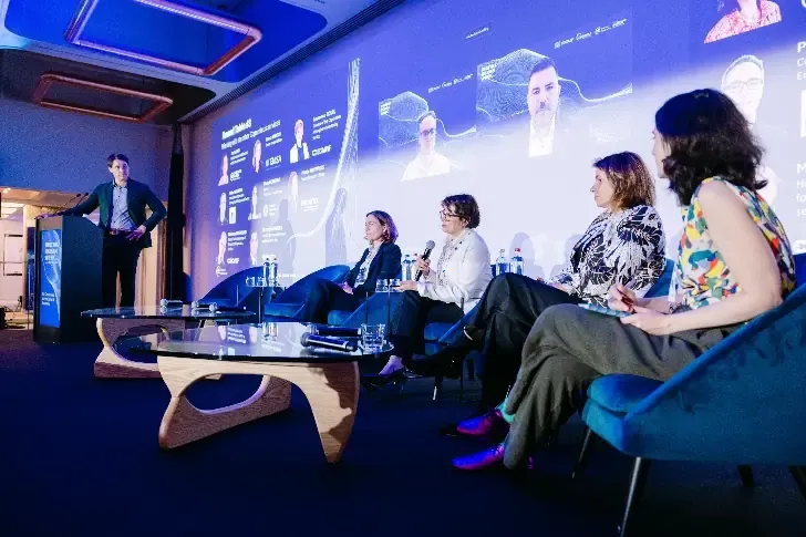 A speaker at a lectern addresses four seated panellists in front of a presentation screen.