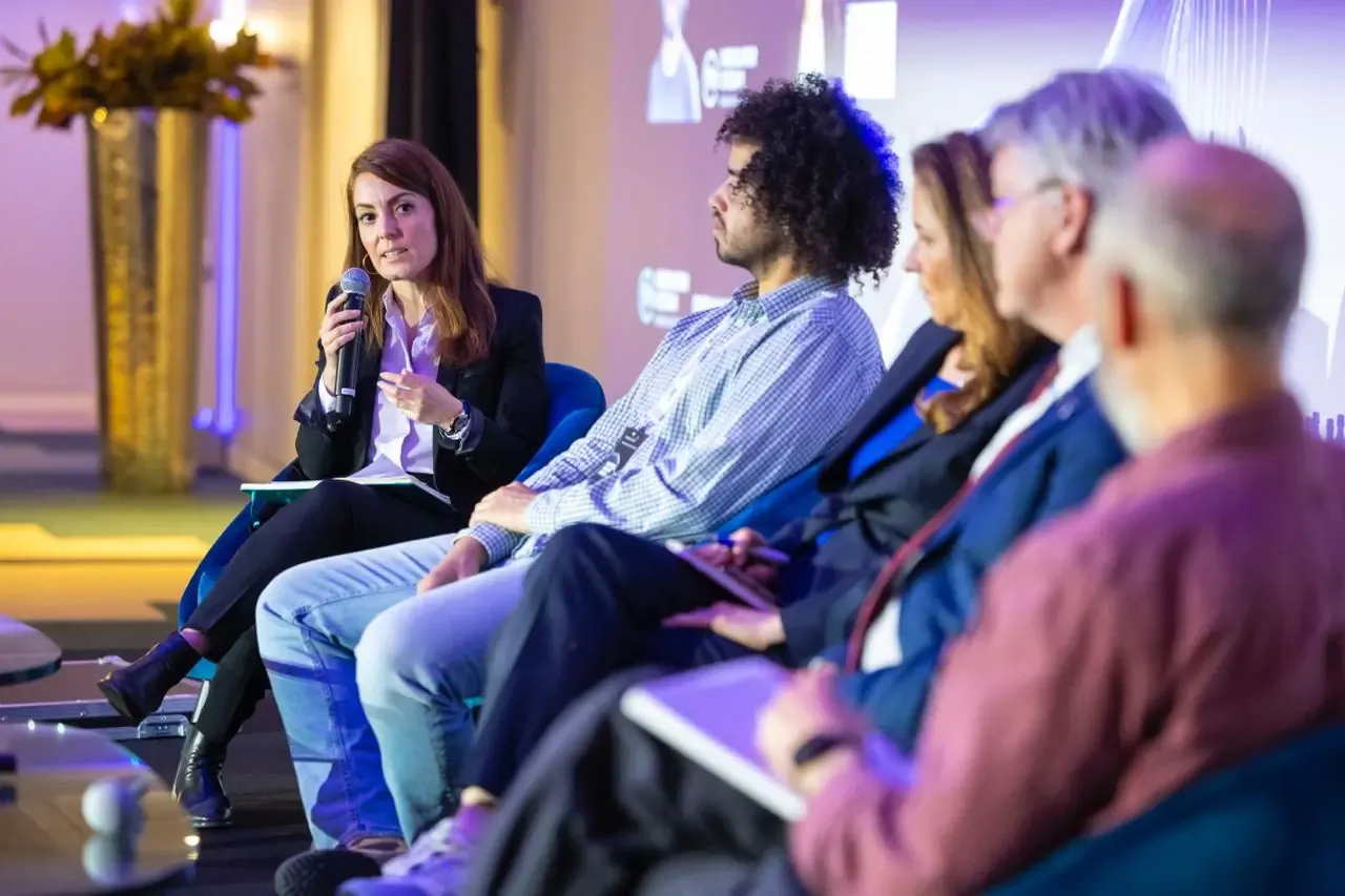 A panel of five speakers seated on a stage at a forum; a woman on the left holds a microphone and speaks while the others listen.