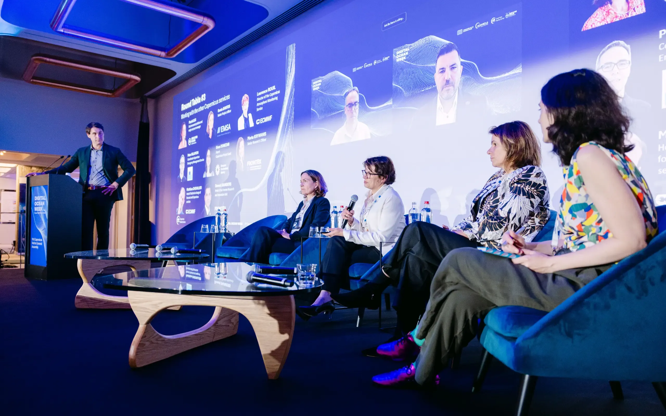 A speaker at a lectern addresses four seated panellists in front of a presentation screen.