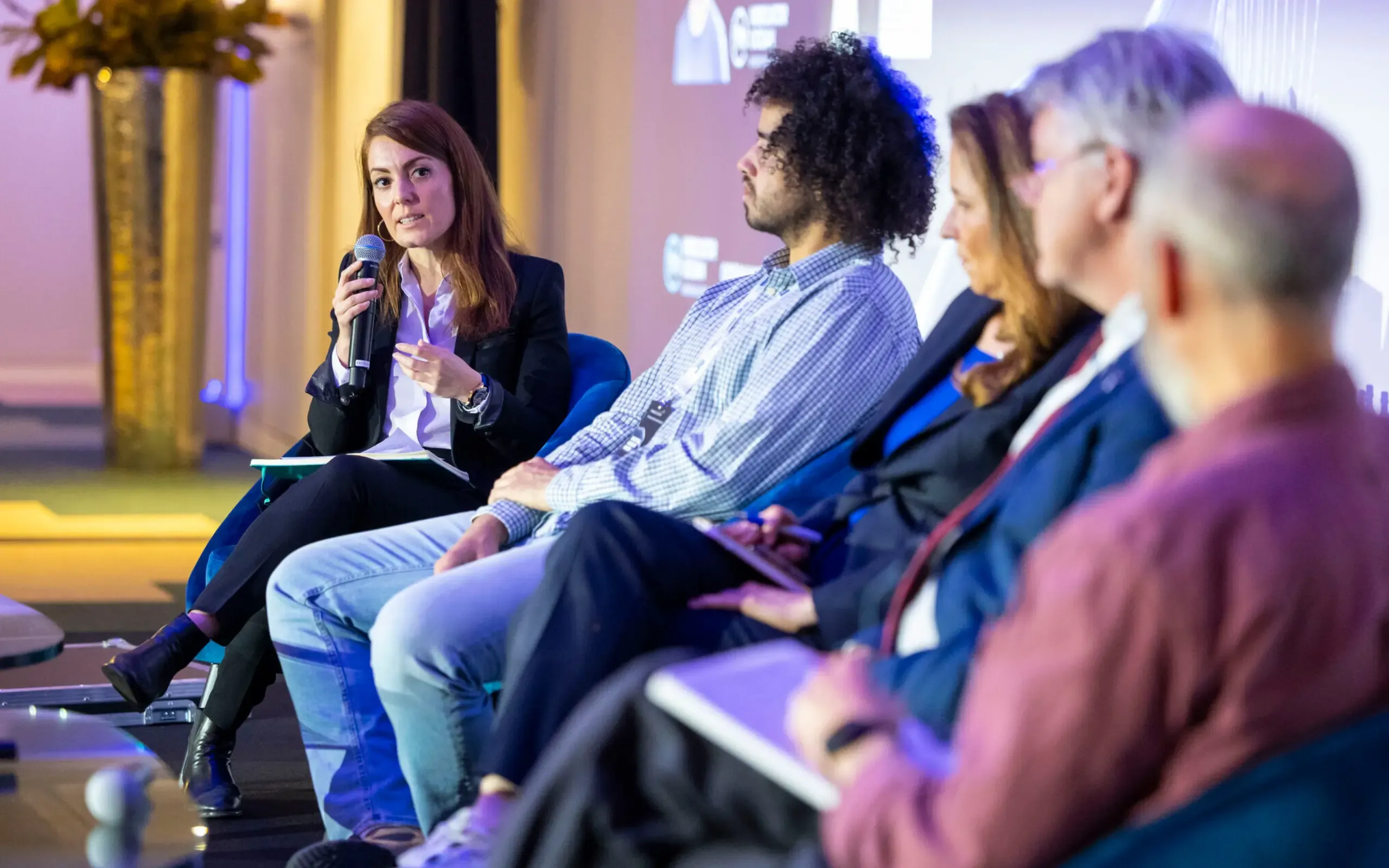 A panel of five speakers seated on a stage at a forum; a woman on the left holds a microphone and speaks while the others listen.
