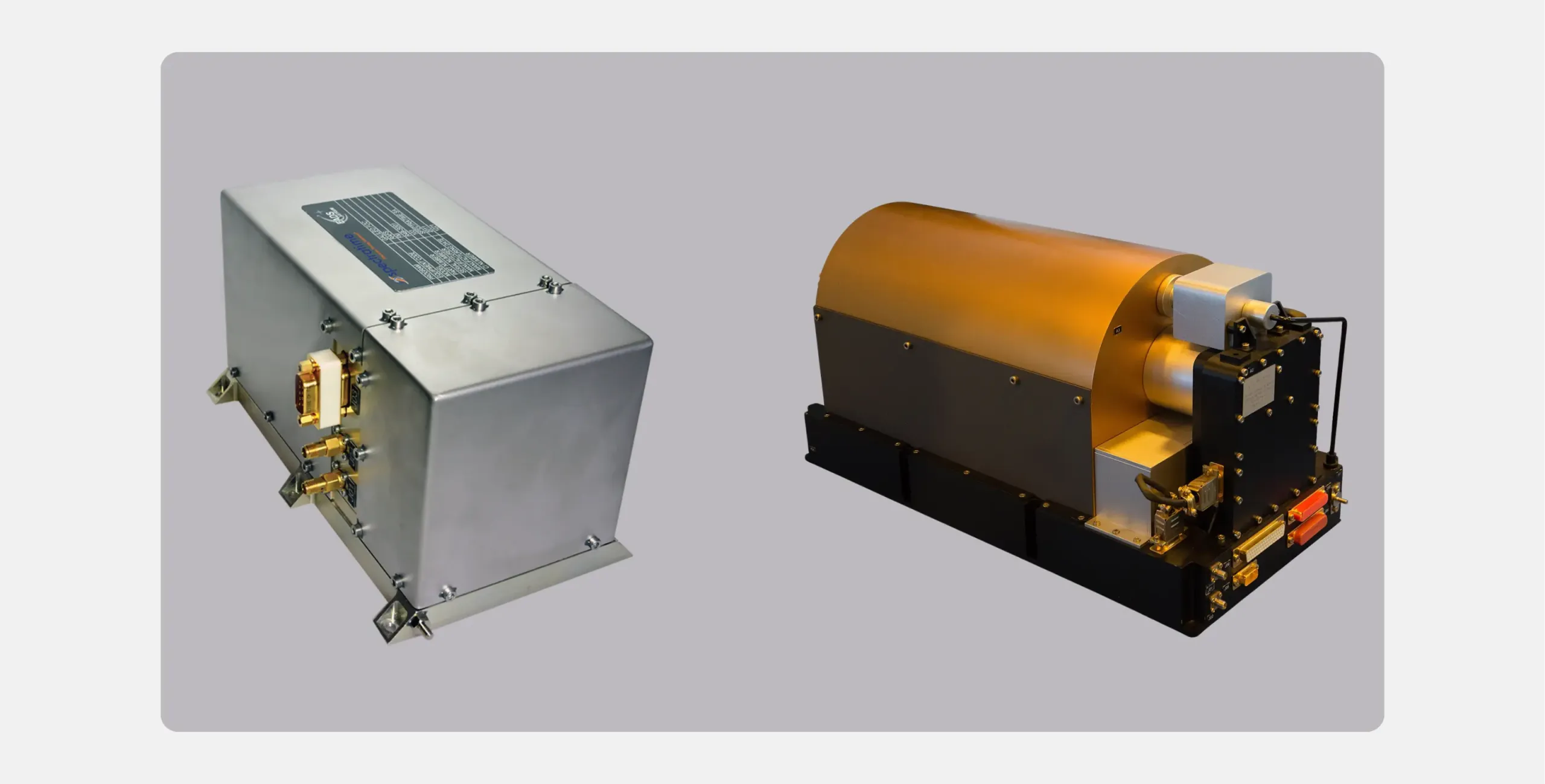 The two types of atomic clock carried on first-generation Galileo satellites, shown side by side. On the left, the rubidium atomic frequency standard: a compact brushed-metal box with a printed label on top and electrical connectors on one end. On the right, the passive hydrogen maser: a larger unit on a black base, with a rounded copper-coloured cylindrical cover and connectors clustered at one end.