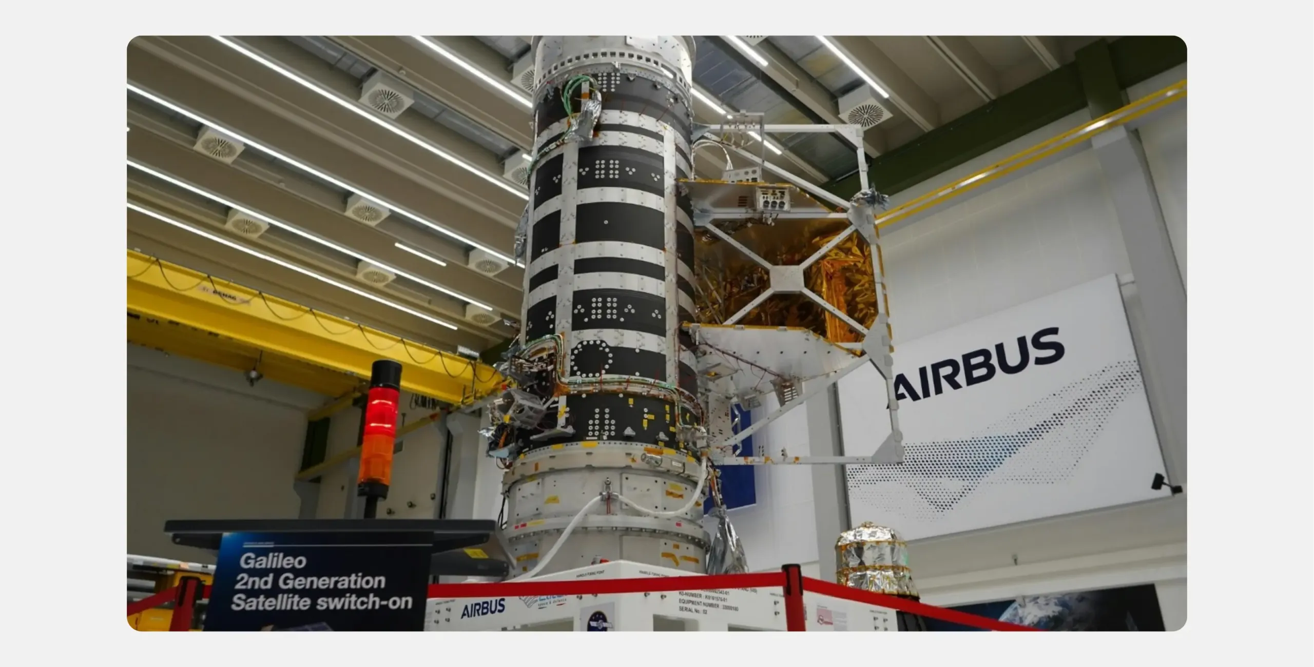 A Galileo Second Generation satellite stands partly assembled in an Airbus cleanroom, its body wrapped in black and white panelling with gold thermal foil visible on one folded side. A sign in the foreground reads "Galileo 2nd Generation Satellite switch-on"