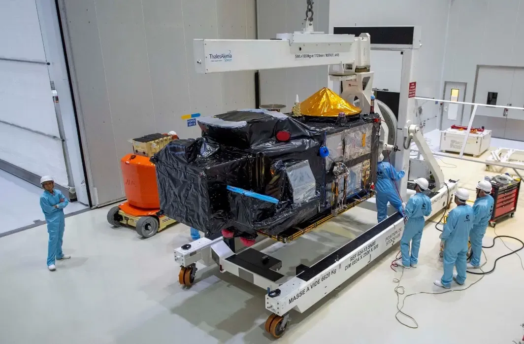 Technicians in blue coveralls and hard hats working on a black foil-wrapped satellite mounted on a transport frame in a clean room.