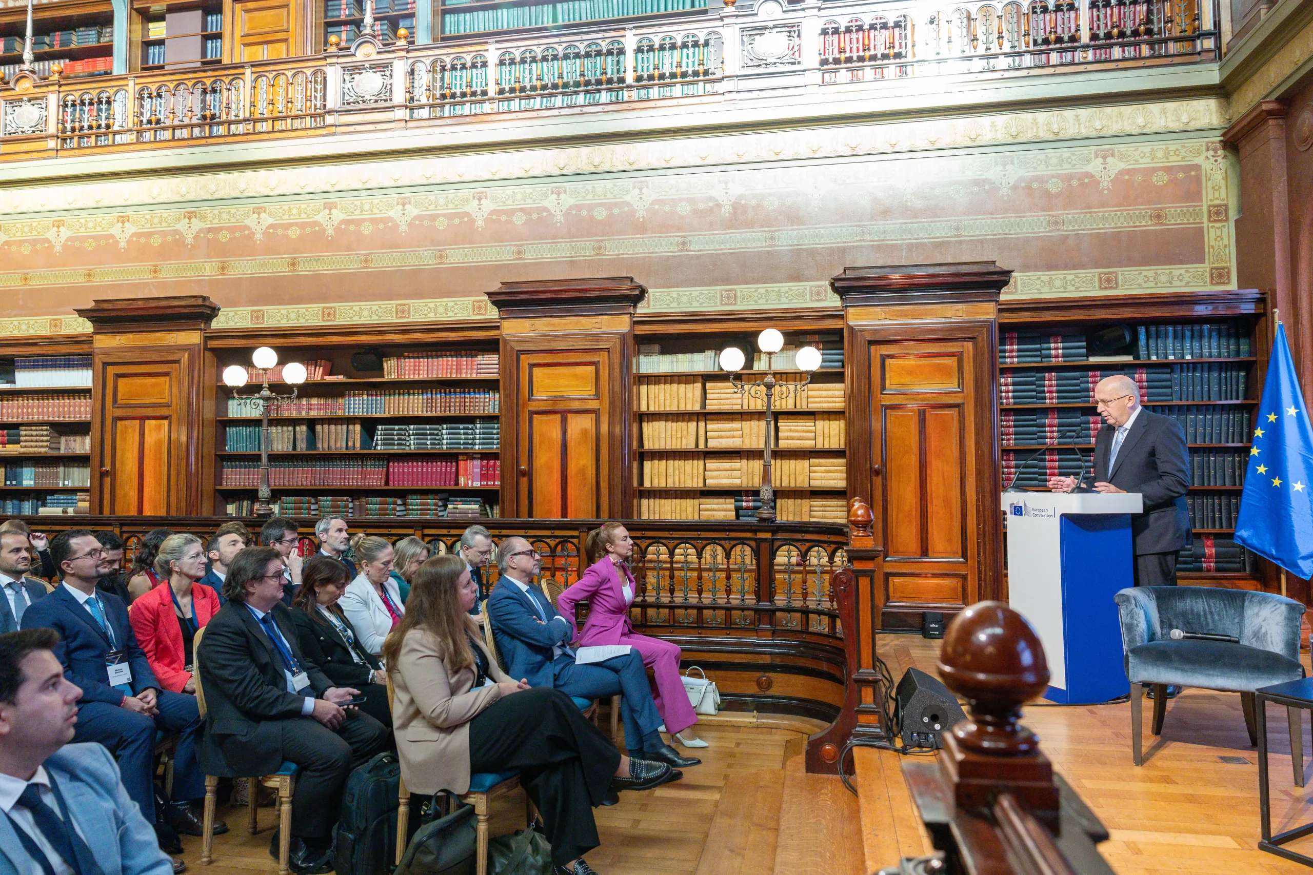 A speaker addresses a seated audience from a European Commission podium beside an EU flag, in the wood-panelled Solvay Library with its galleried bookshelves.