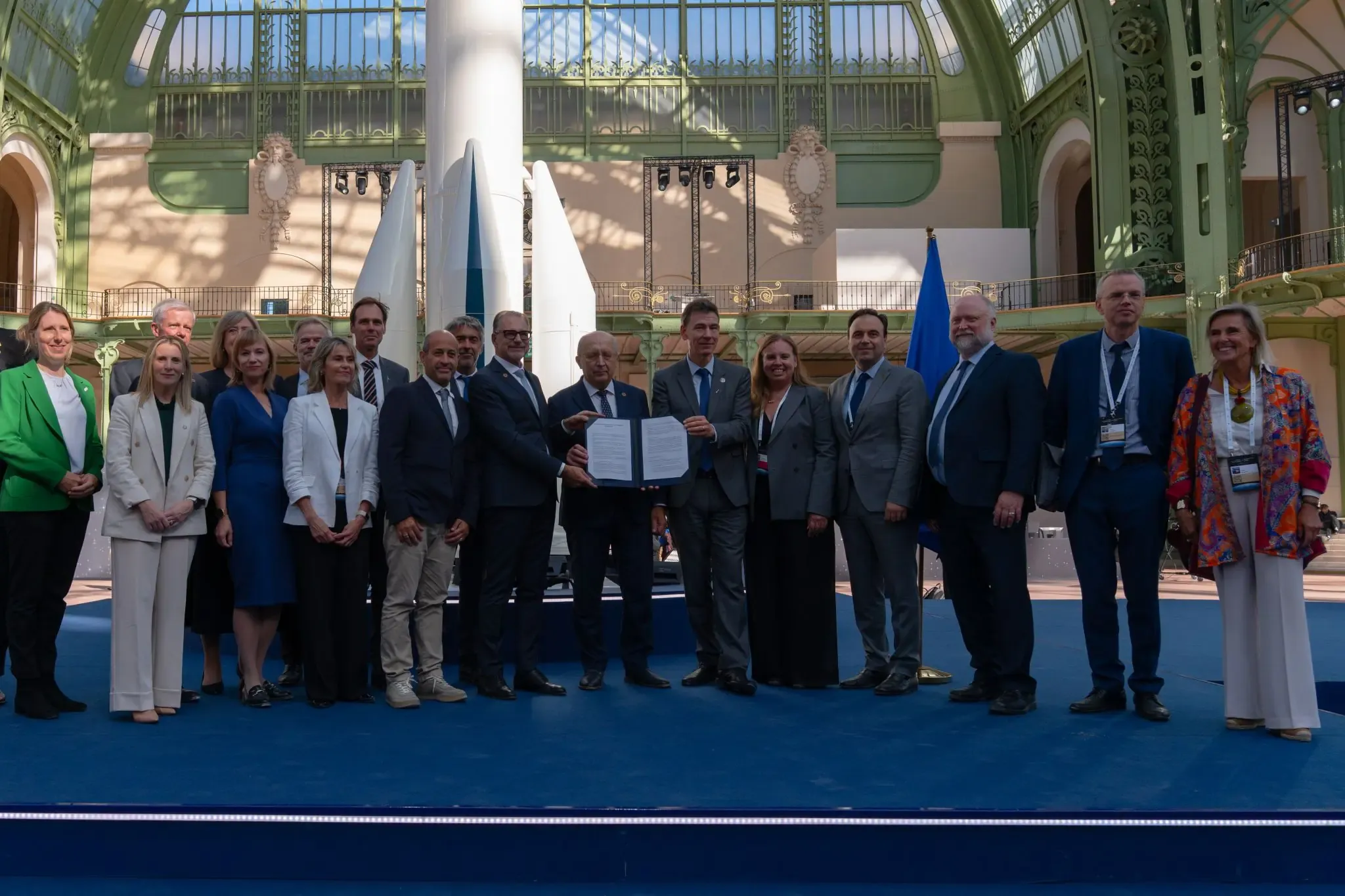A group of delegates in business attire stand in a row on a blue stage. Two people at the centre hold open a signed declaration towards the camera. A European Union flag stands to their right. Behind them are white and teal scale models of space launchers, beneath the arched glass-and-ironwork dome of the Grand Palais in Paris.