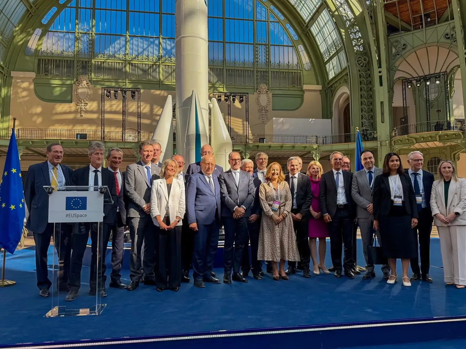 A group of delegates in business attire stand together on a blue stage behind a podium bearing the EU emblem and the hashtag #EUSpace. Two European Union flags flank the group. Behind them are white and teal scale models of space launchers, set beneath the large arched glass-and-ironwork dome of the Grand Palais in Paris.