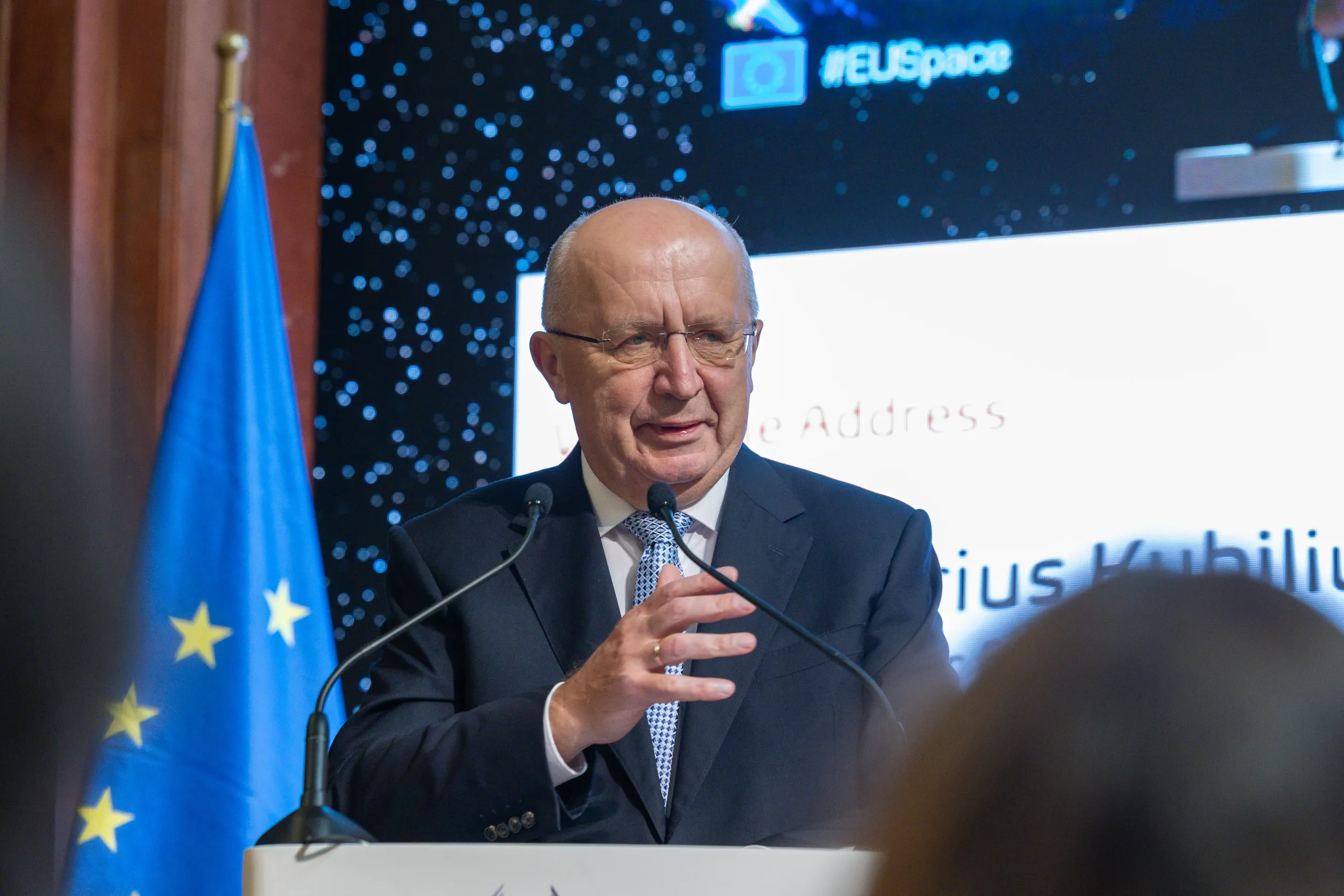 Andrius Kubilius, European Commissioner for Defence and Space, speaking at a podium during the welcome address, with an EU flag behind him.