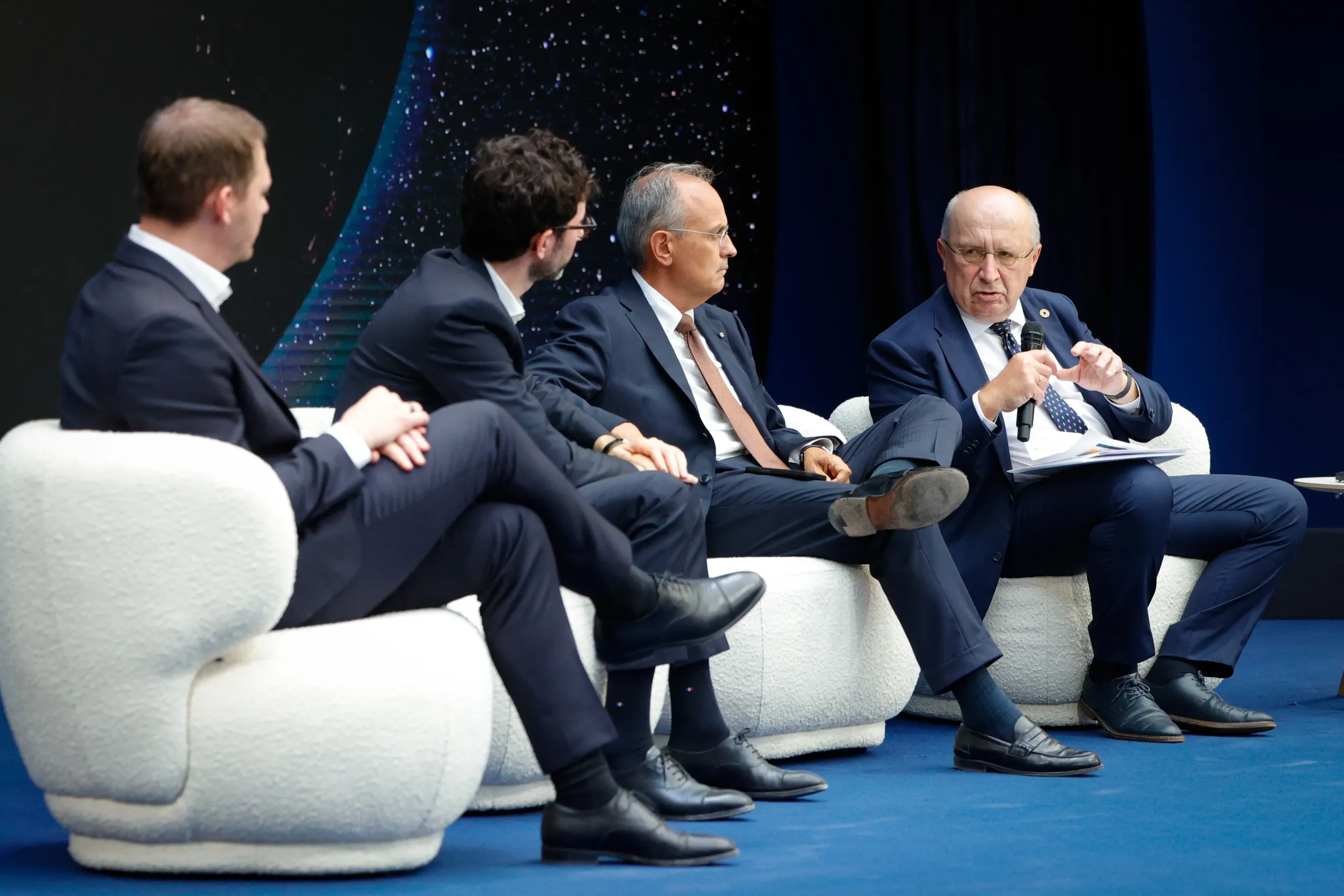 Four men in dark suits sit in white armchairs on a blue stage during a panel discussion, against a dark starfield backdrop. The man on the right speaks into a handheld microphone while holding papers; the others listen.