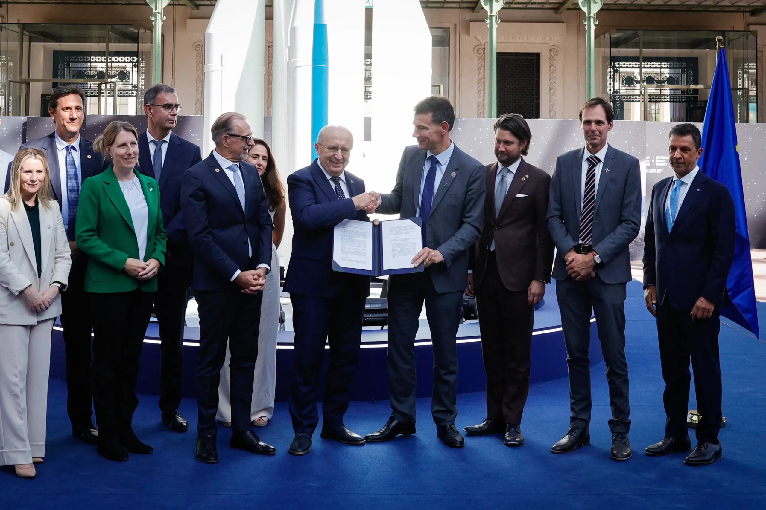 A group of people in business dress stand in a row for a photograph. Two men at the centre shake hands while jointly holding an open signed document. A model rocket and a European Union flag stand behind them.