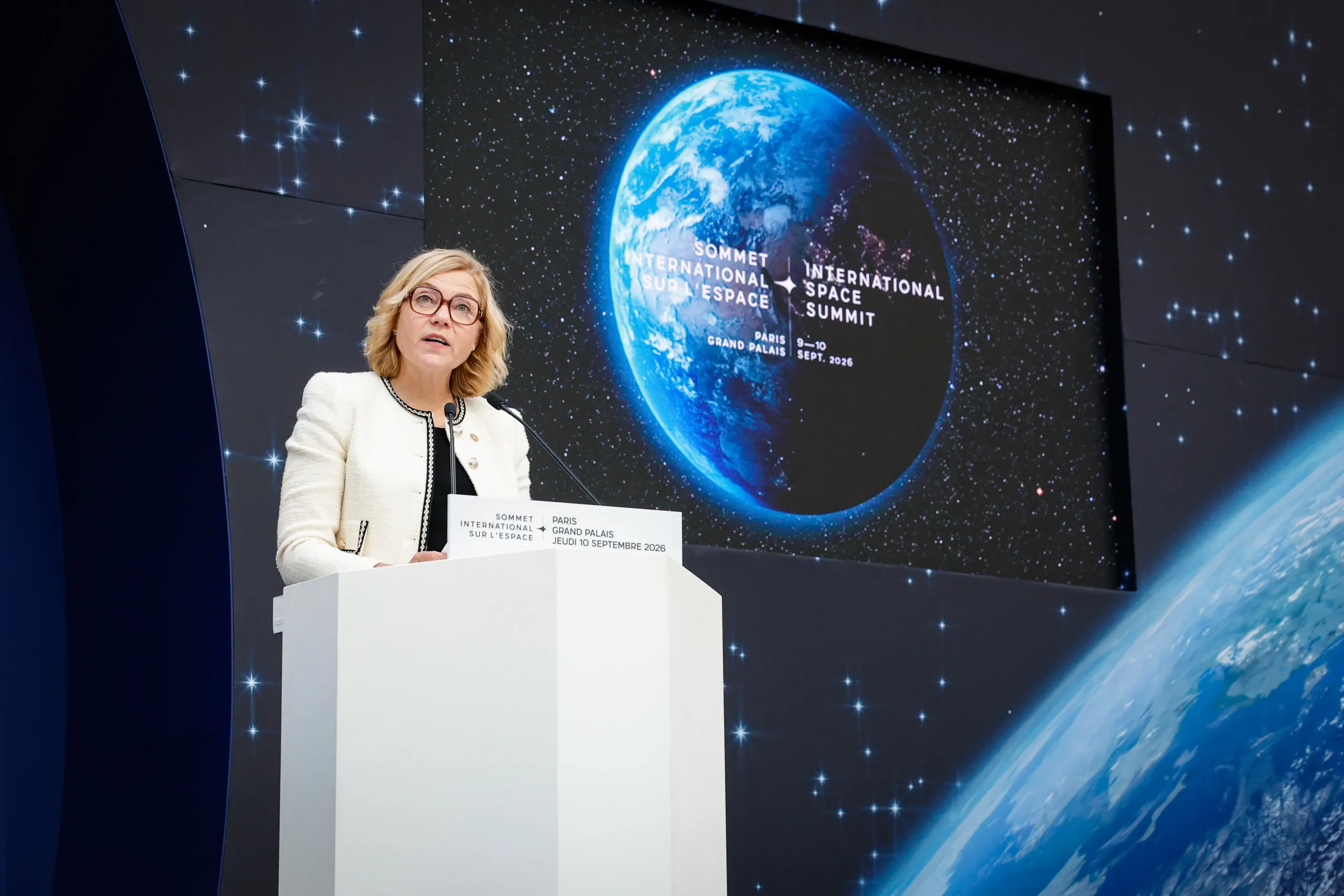 A woman in a white jacket and red glasses speaks at a white lectern, in front of a large screen showing Earth and the International Space Summit logo, dated 9–10 September 2026, Paris, Grand Palais.