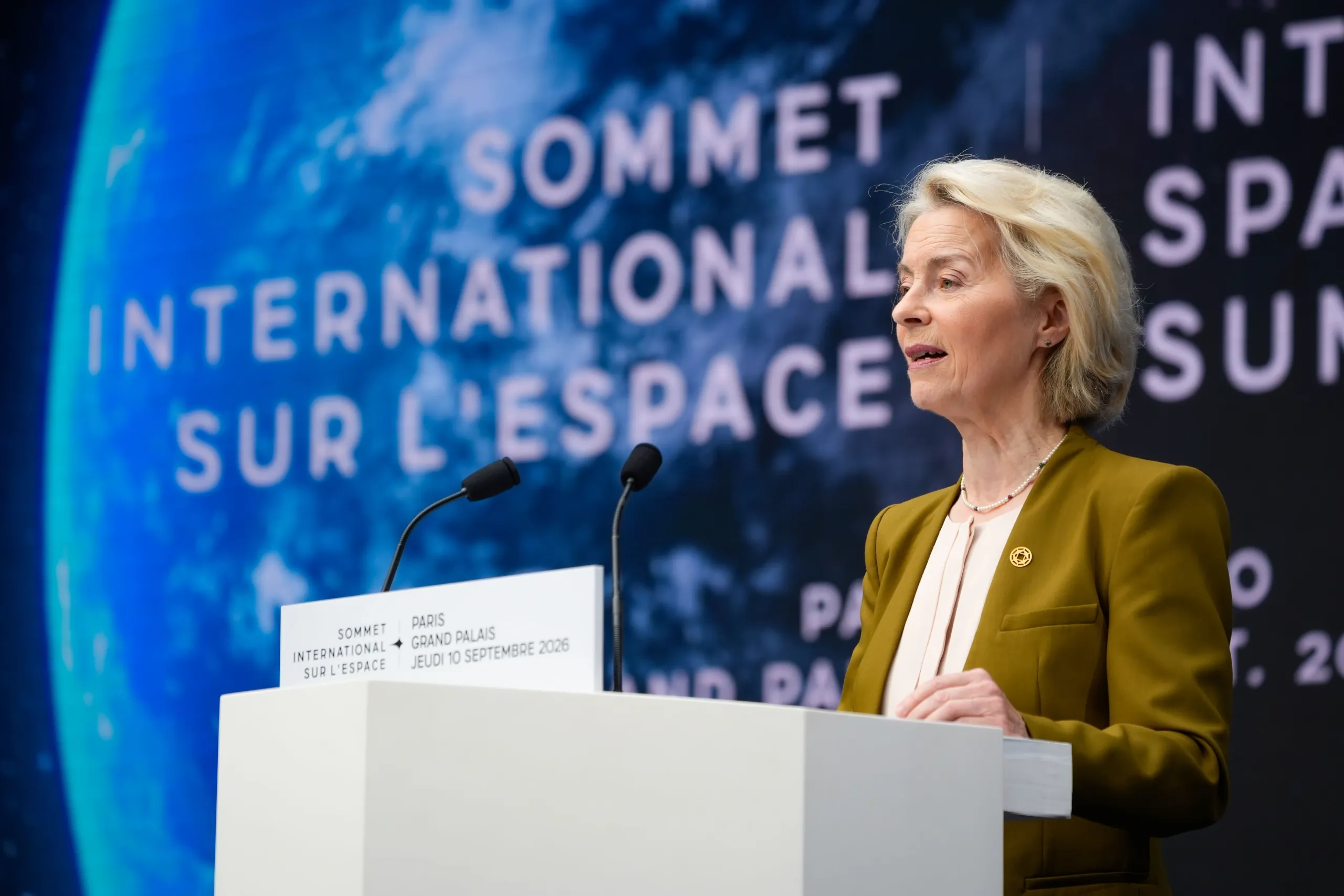 A woman in an olive-green blazer speaks at a white lectern into two microphones, in front of a large screen reading 'Sommet International sur l'Espace' over an image of Earth. The lectern reads 'Paris, Grand Palais, Jeudi 10 Septembre 2026'.