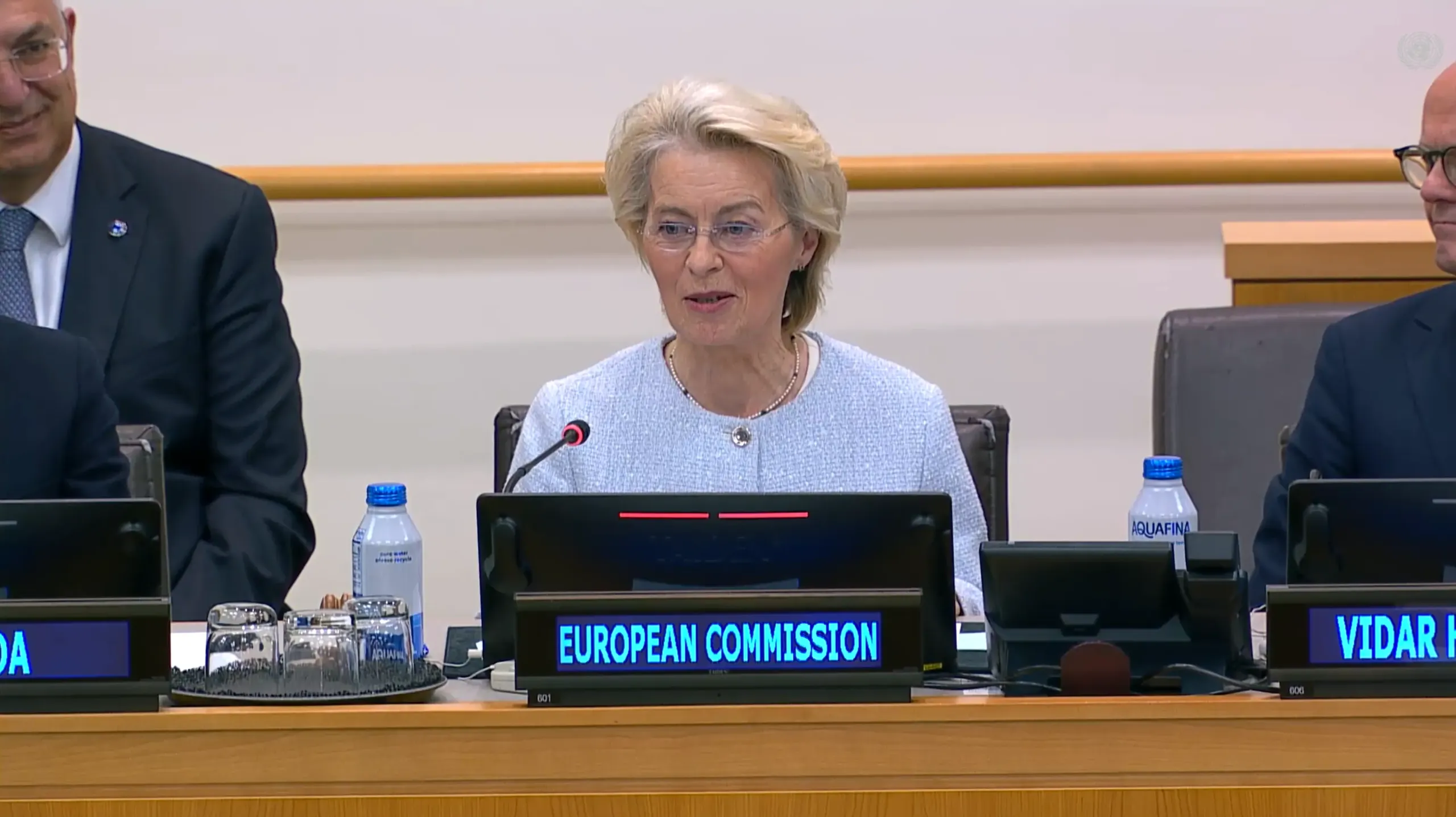 Ursula von der Leyen speaks into a microphone at a conference table behind a nameplate reading "European Commission", with delegates seated on either side.