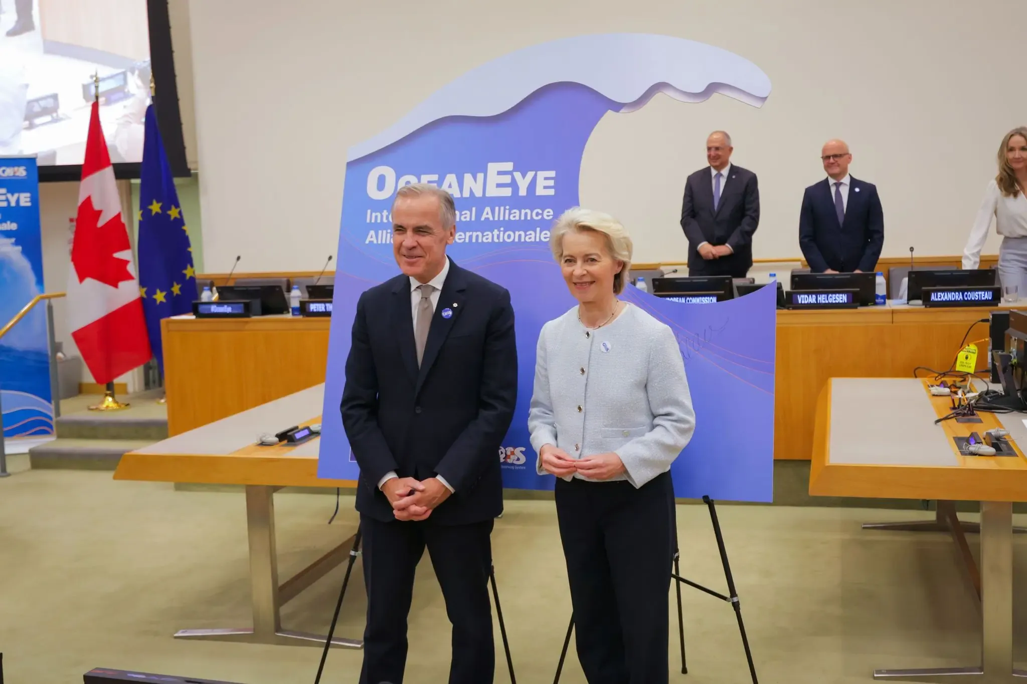 Mark Carney and Ursula von der Leyen stand in front of a wave-shaped OceanEye International Alliance sign, with Canadian and EU flags to the left and delegates standing at the conference tables behind them.