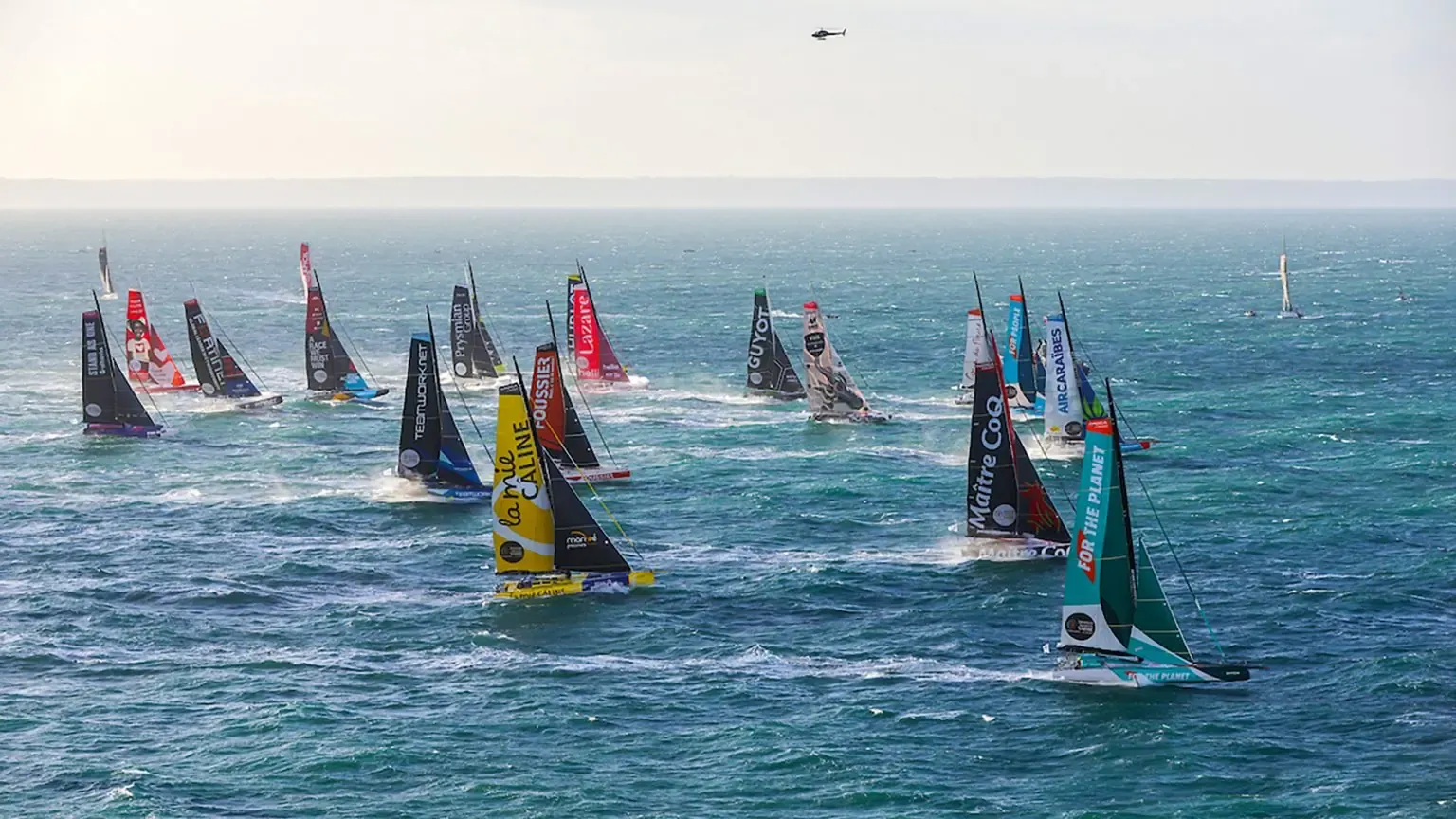 Aerial view of several Vendée Globe racing yachts with dark and coloured sails crossing calm blue sea, with support boats in the distance.