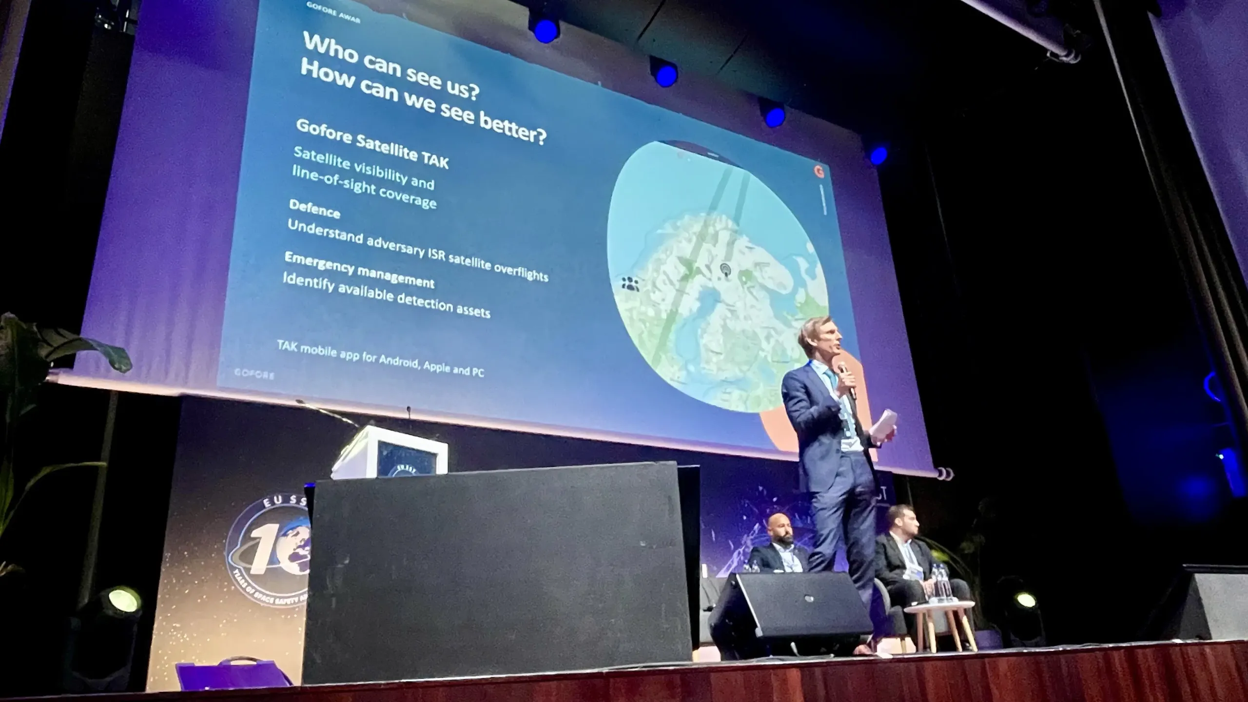 A speaker presents on stage at the EU SST 10-year anniversary event in front of a slide from the Finnish company Gofore. The slide is titled “Who can see us? How can we see better?” and shows the company’s satellite visibility application over a map of northern Europe. Two other speakers sit in the background.
