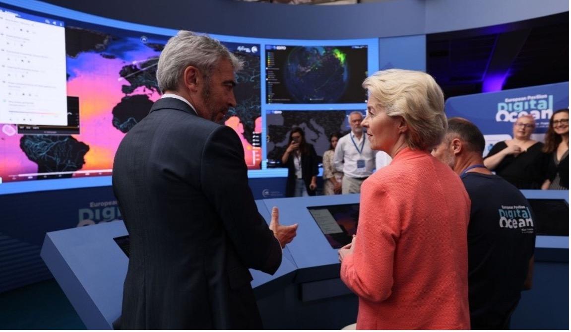 Two individuals discuss in front of large screens displaying colorful maps and data, with a group of people observing in the background.