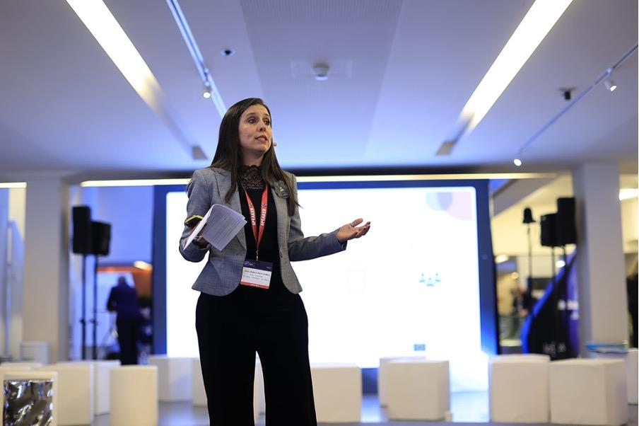 A person in a blazer speaks on stage holding papers, with a large screen and audience seating visible in the background.
