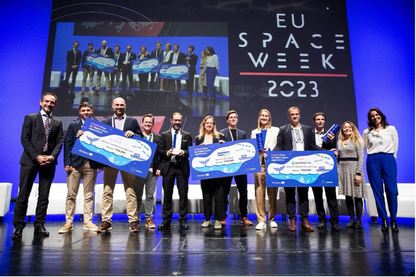 The image shows the winners of the CASSINI Maritime Prize at EU Space Week 2023 in Seville. SciDrones, EOMAP, and Geomatys representatives stand on stage holding large ceremonial checks and trophies. They are surrounded by officials and other participa...