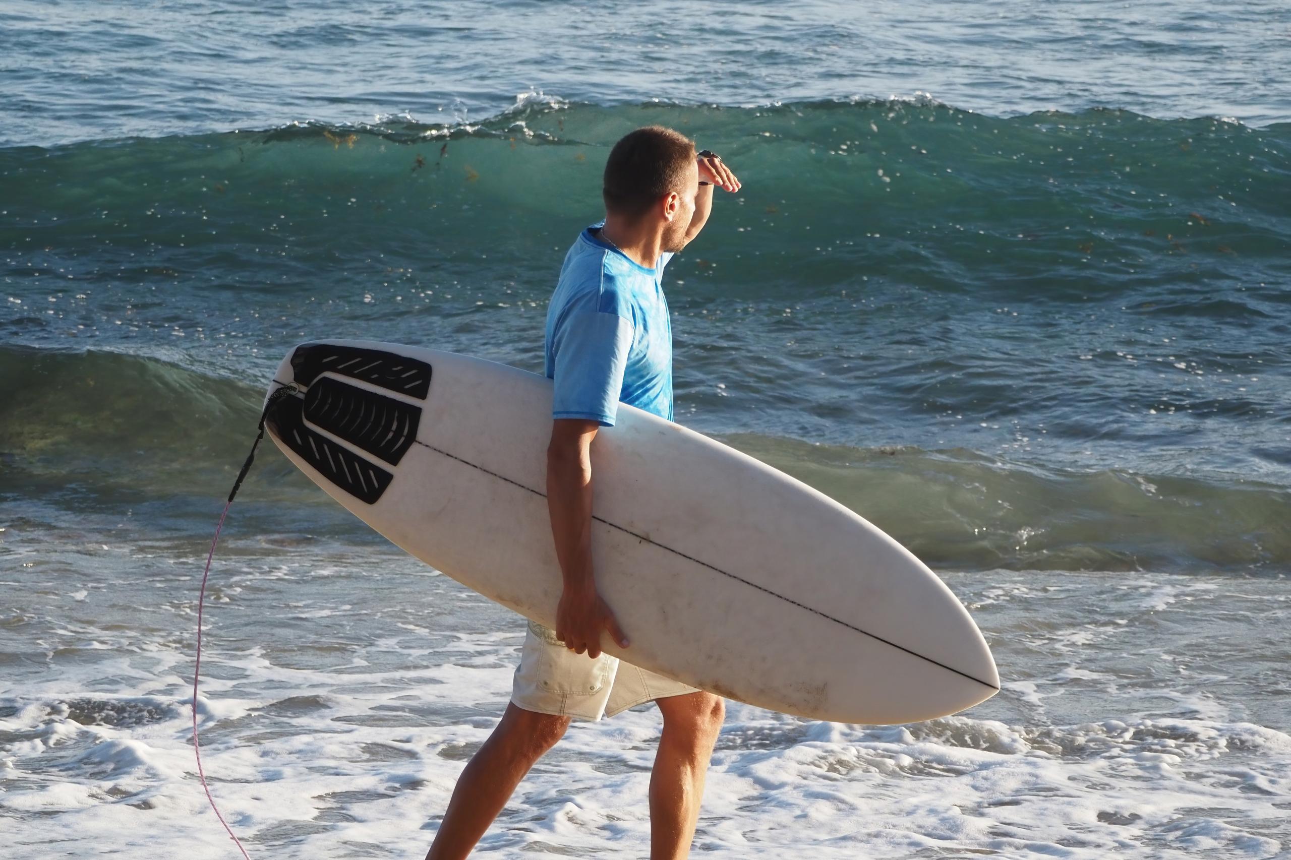 The image shows a man standing on the shoreline, holding a surfboard under his arm. He is wearing a blue shirt and white shorts, looking out towards the ocean, shading his eyes with his hand. The waves are gently crashing on the beach, and the water is...