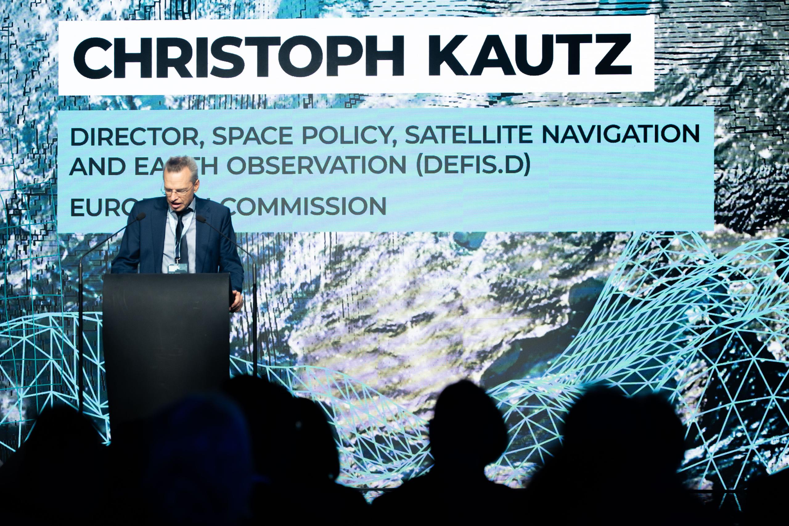 Christoph Kautz, Director for Space Policy, Satellite Navigation and Earth Observation (DEFIS.D) at the European Commission, speaks at a podium during the “10 Years of Copernicus at Frontex” event. Behind him, a large screen displays his name, title, a...