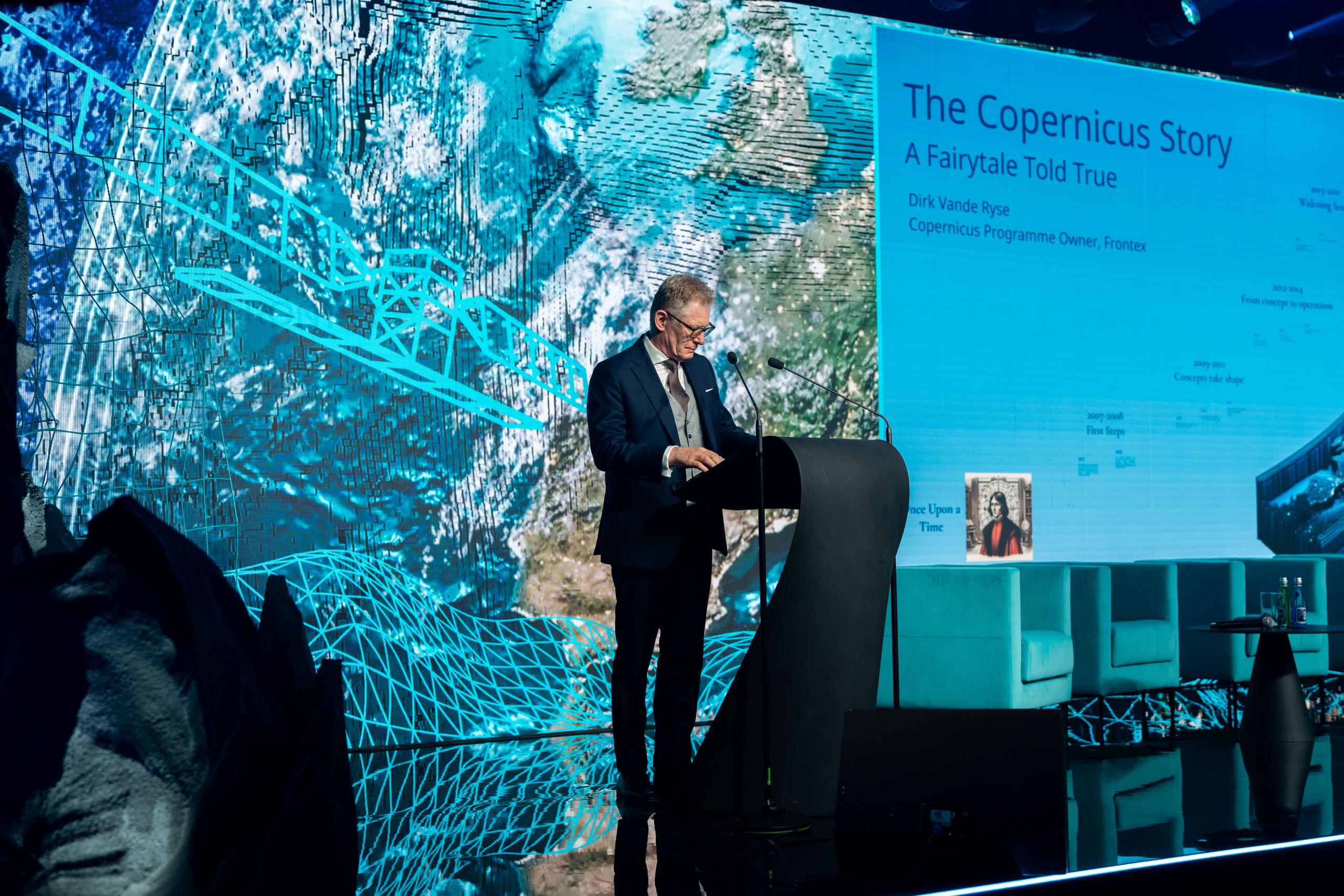 A man in a suit stands at a podium on stage, giving a presentation titled “The Copernicus Story: A Fairytale Told True.” The background screen shows Earth from space with digital blue grid overlays and text about Copernicus.