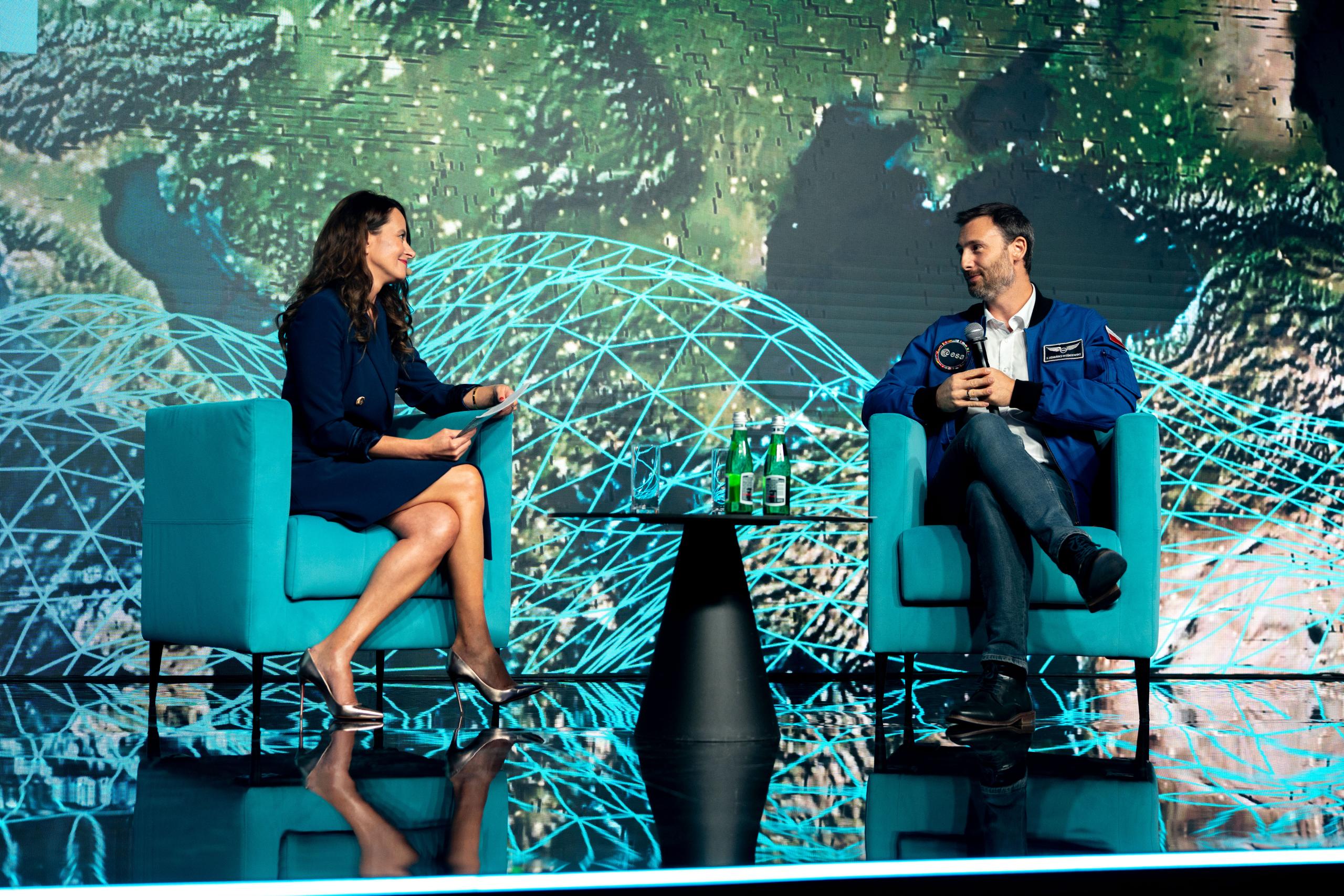 A woman interviewer and a Astronaut Sławosz Uznański-Wiśniewski in a blue jacket sit on turquoise armchairs on stage, engaged in a discussion. A small table with water bottles and glasses is between them, with a large digital backdrop showing Earth and...