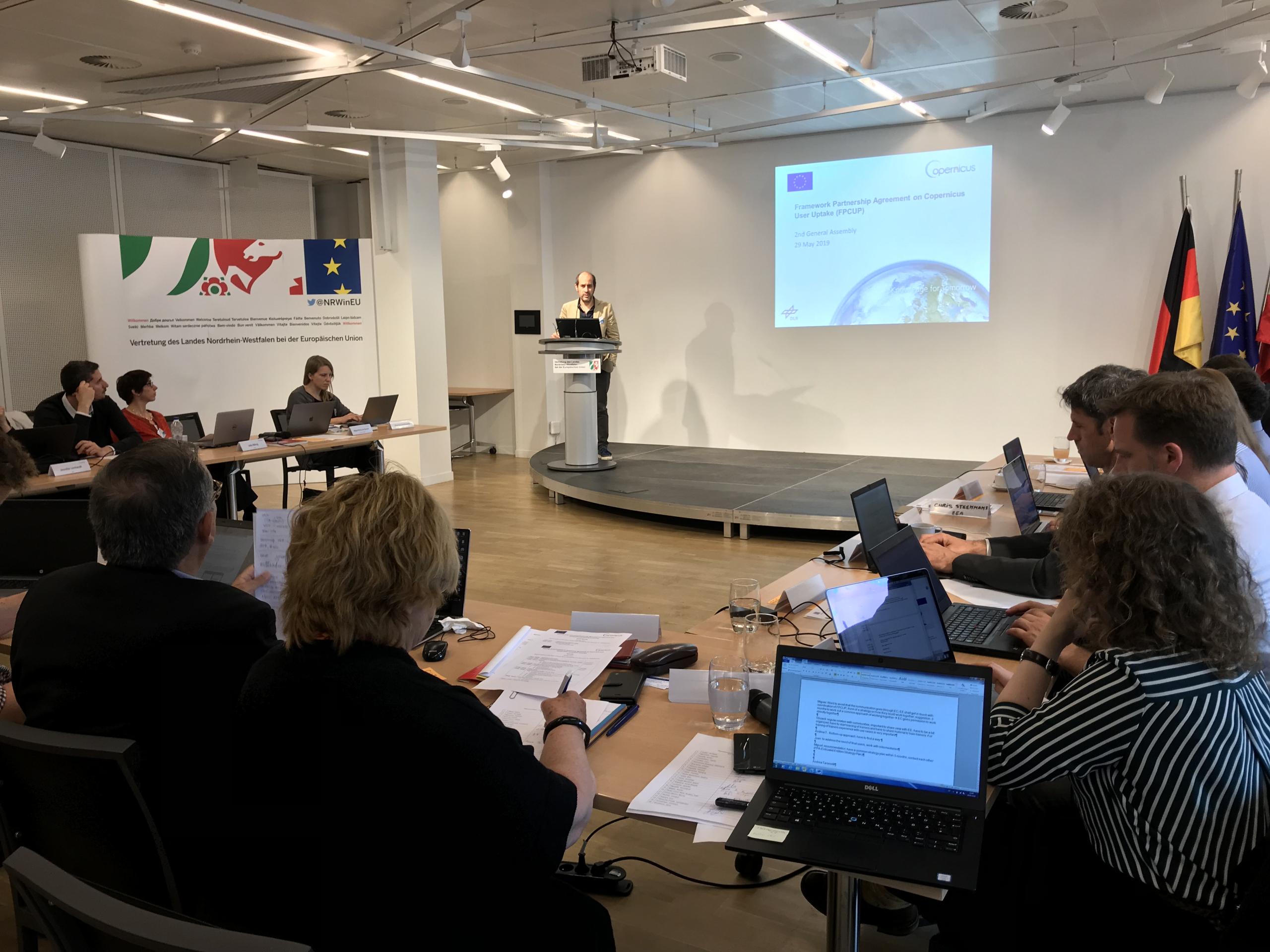 A man stands at a podium delivering a presentation in a conference room. The presentation slide behind him is titled “Framework Partnership Agreement on Copernicus User Uptake (FPCUP)” dated 29 May 2019, with the EU and Copernicus logos. Around the roo...