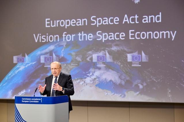 Commissioner for Defence and Space, Andrius Kubilius standing at a podium with a blue banner reading 'Commission européenne European Commission.' Behind him is a large screen displaying an image of Earth from space with European Union flags and the tex...