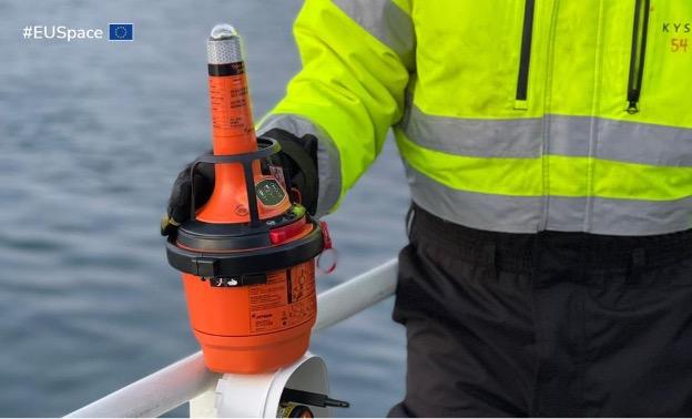 The image shows a person wearing a high-visibility yellow jacket with reflective stripes, holding an orange emergency beacon. The person is standing on a boat, with the ocean visible in the background. The beacon is equipped with various dials and a li...