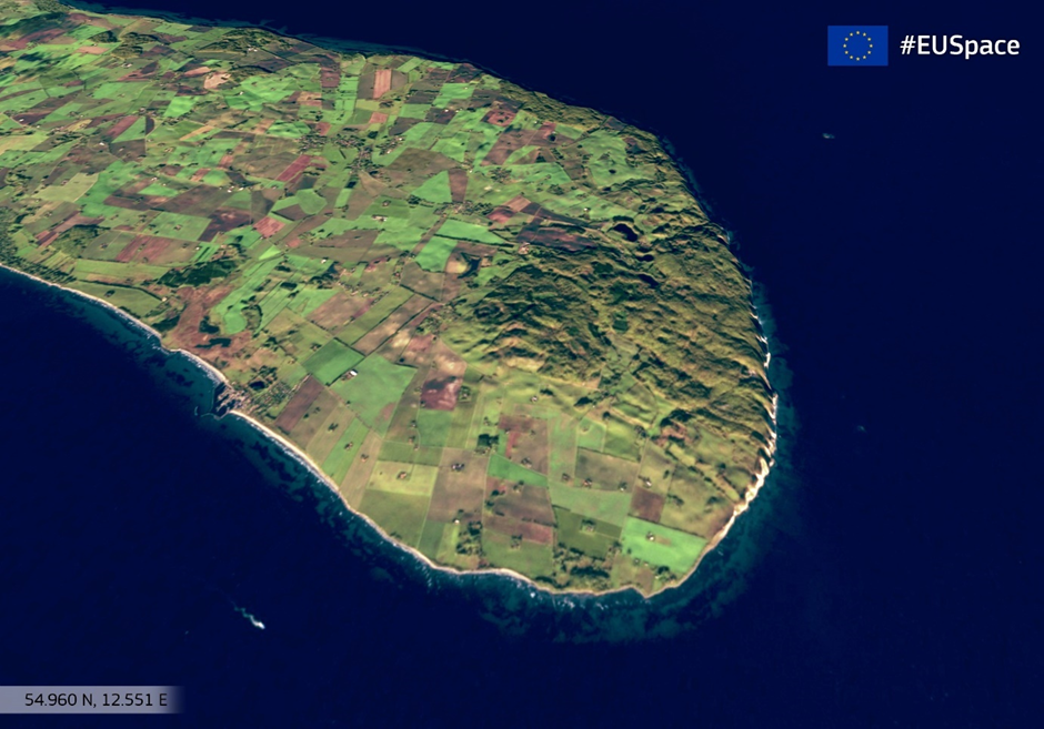 This image shows a satellite view of a coastal region, likely captured by the Copernicus Sentinel-2 satellite. The image features a landscape with a patchwork of green fields extending to the coastline, bordered by the deep blue sea. The high-resolutio...