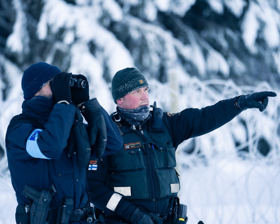 Two officers in winter gear observing a snowy landscape; one uses binoculars, the other points ahead, with a barbed wire fence in the background.