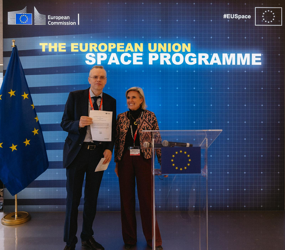 A man in a dark suit and glasses holds up a signed document while standing next to a smiling woman in a patterned jacket and dark trousers. They are in front of a blue backdrop displaying the text 'THE EUROPEAN UNION SPACE PROGRAMME' along with the Eur...