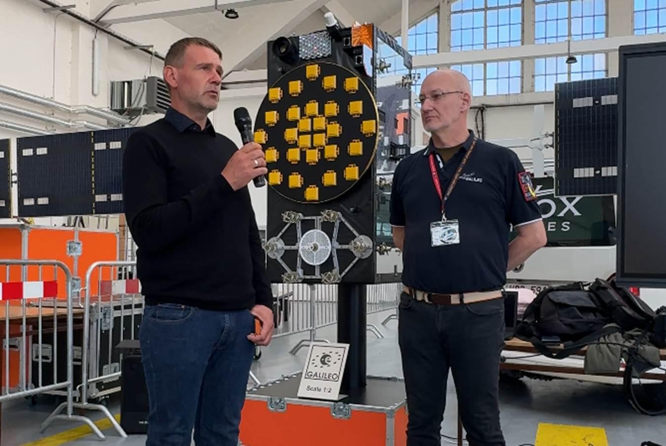 the image shows two people talking in front of a Galileo satellite model