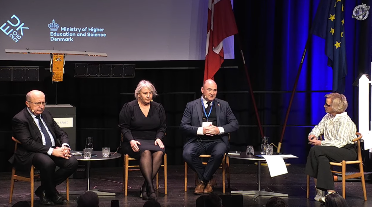 Four people are seated on stage during a panel discussion. Behind them are the flags of Denmark and the European Union, along with a banner for the Ministry of Higher Education and Science of Denmark.